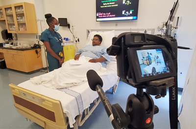 A camera in the foreground films a doctor and standardized patient for a video simulation.